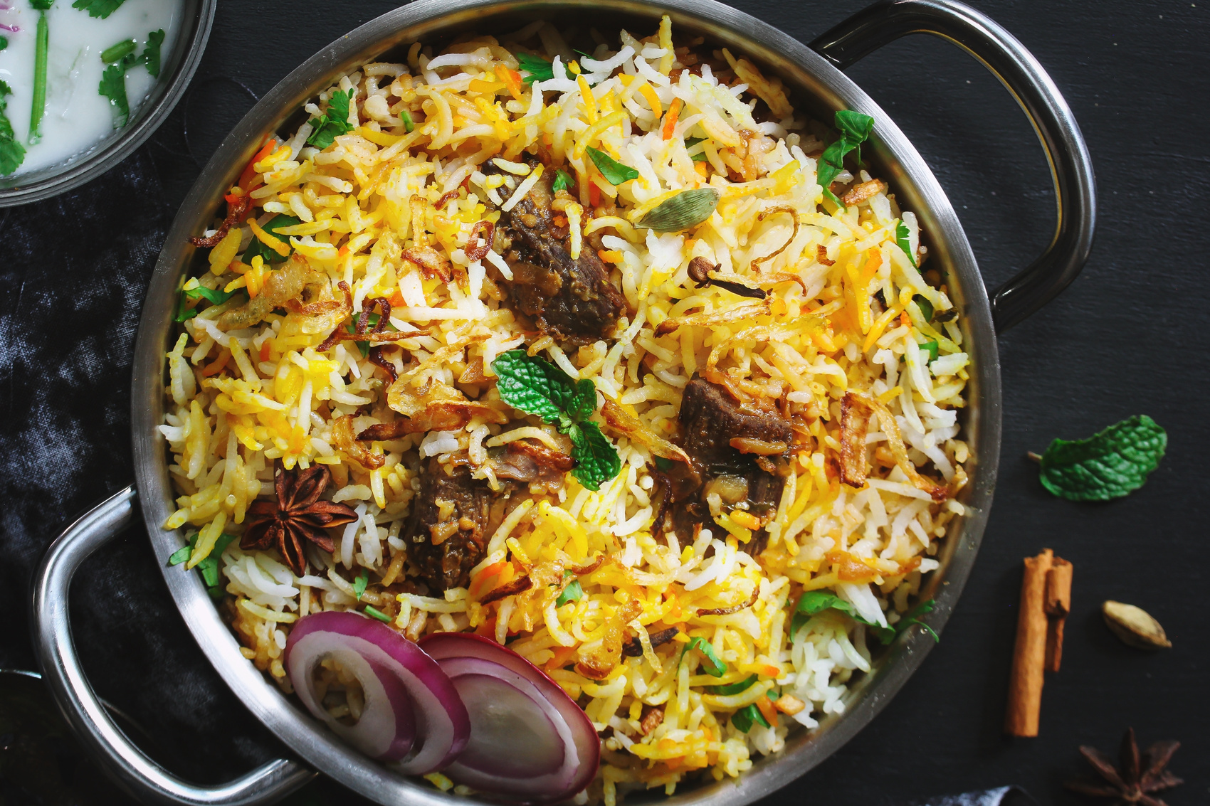 delicious hotel daawat colourful mutton biryani beautifully presented with fried onions, raw onions, star anise, cloves and in a pan on a black background